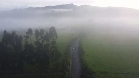 Drohnenflug-über-Der-Autostraße-Im-Dichten-Nebel-Auf-Einer-Großen-Grünlandfläche-Mit-Hügeln-Im-Hintergrund