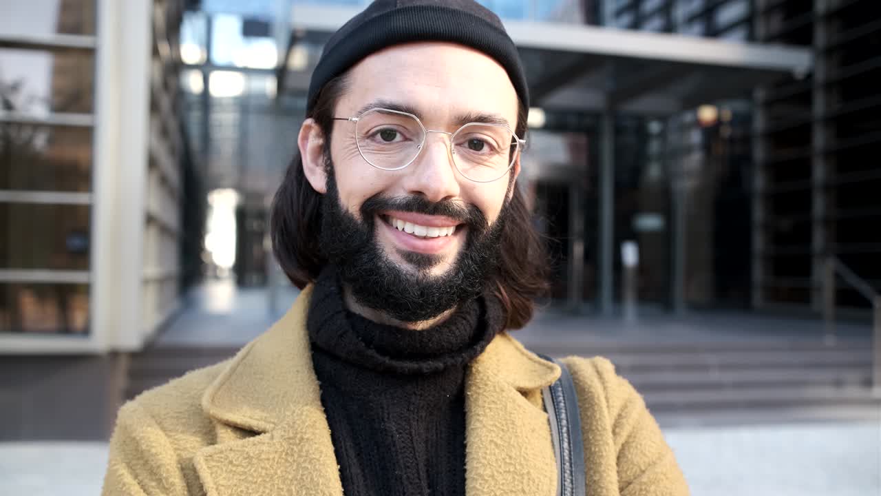 joven mirando a la cámara con una sonrisa en la cara mientras está de pie contra un edificio de oficinas.