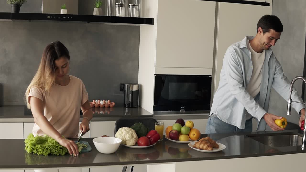 Cheerful young family couple cutting fresh vegetable salad, having fun cooking together im modern loft interior kitchen, happy husband helping prepare healthy meal - washing yellow peper in sink