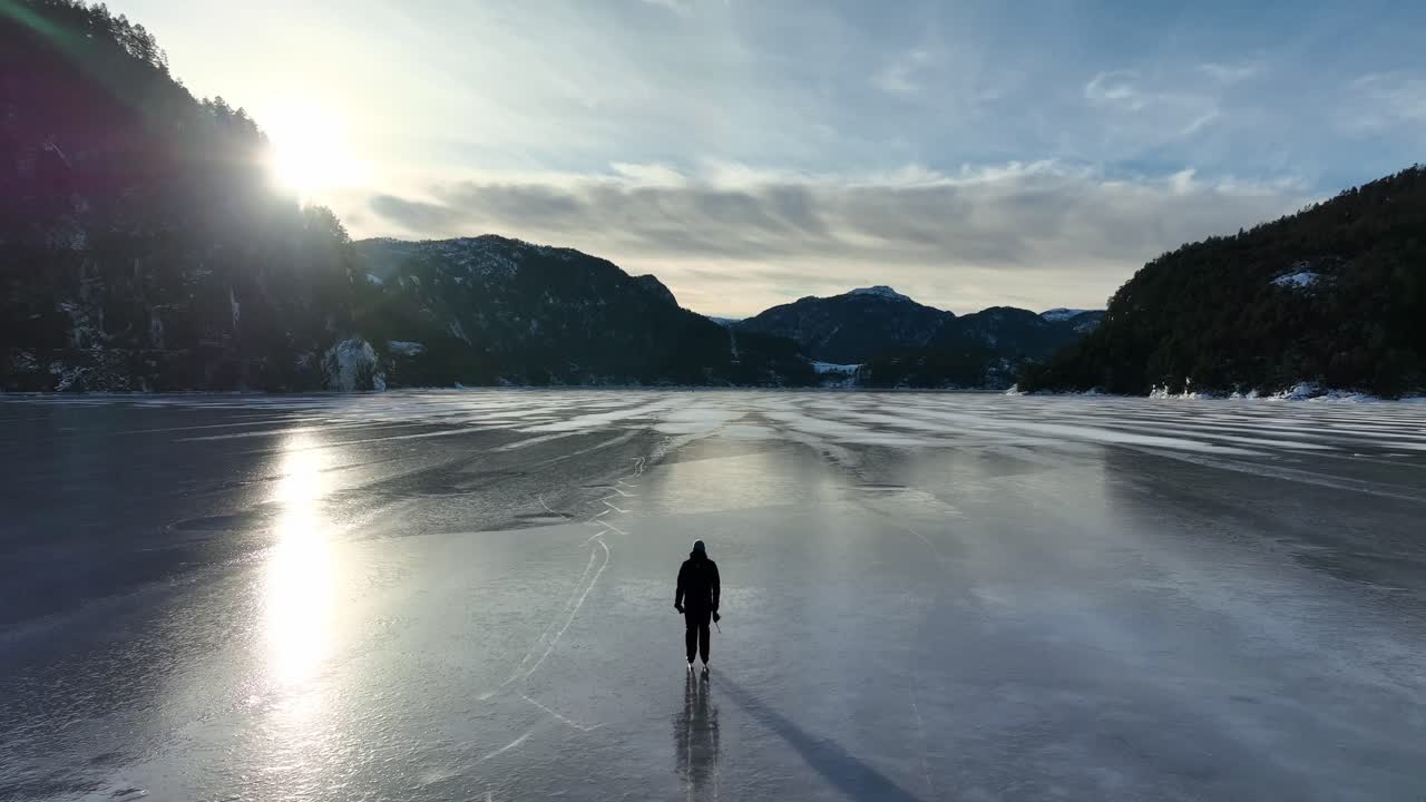 Aerial glide crosses frozen Eidsfjord toward sunset as skater enters frame then moves away across vast ice in winter Norway