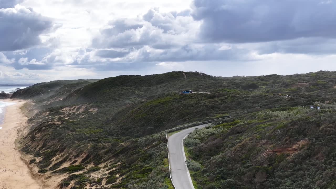 Drone pans above winding coastal road, sandy beach, and rugged cliffs under dramatic cloudy sky