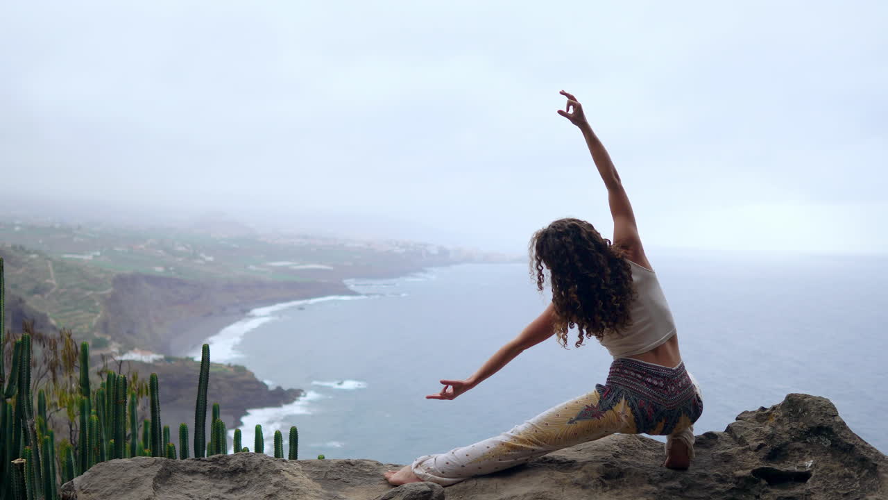 A woman sits on a cliff's edge, striking a warrior pose, raising her arms and inhaling the sea air as she immerses herself in yoga by the ocean