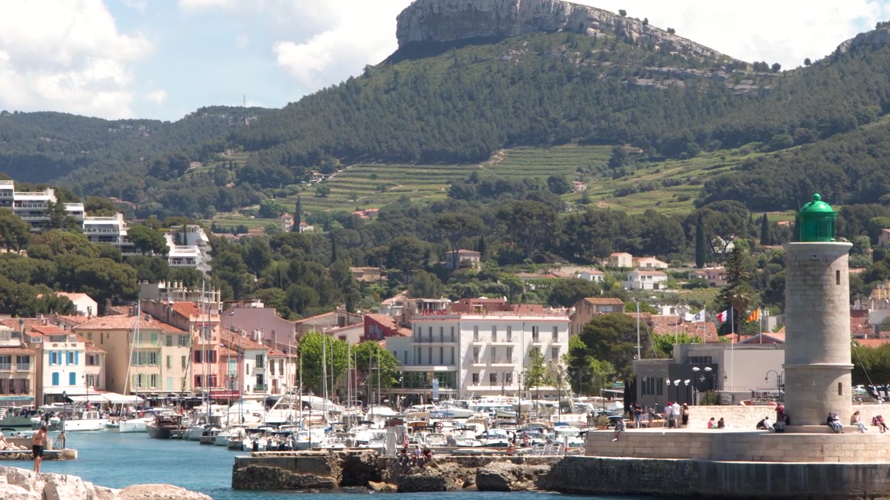 Ampia inquadratura statica di un porto soleggiato con barche, un faro e uno sfondo montuoso.