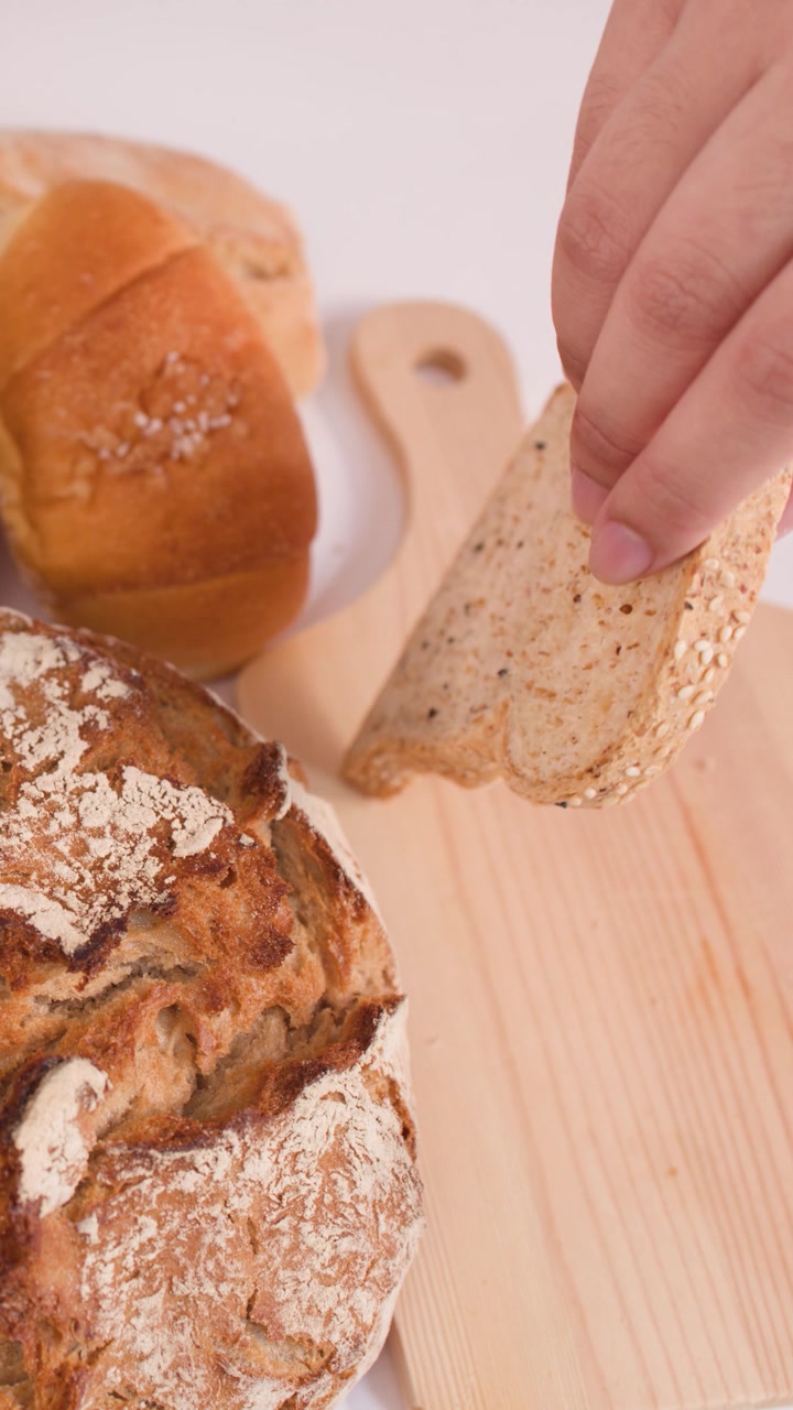 Person places fresh multigrain bread slice onto wooden board beside sourdough and artisan loaves.