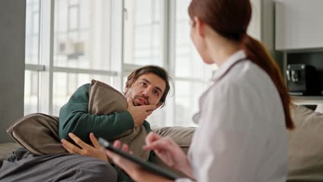 a brunette man with stubble in a green jacket tells a brunette girl doctor in a medical gown about his problems while sitting on the sofa and hugs in a modern apartment during a home examination