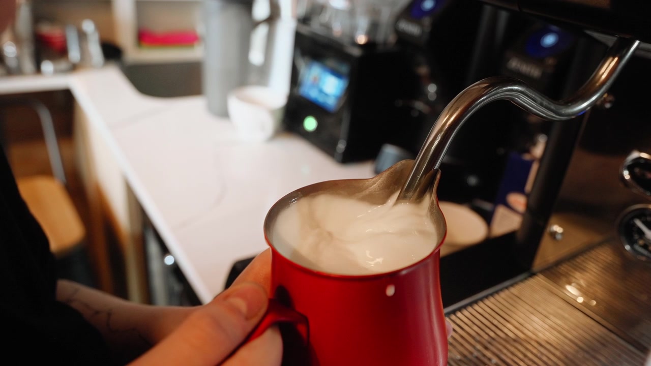 Primo piano di un barista che monta il latte in una lattiera per un cappuccino, all'interno di una caffetteria, al rallentatore.