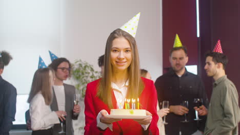 Retrato-De-Una-Hermosa-Mujer-Sosteniendo-Un-Pastel-De-Cumpleaños,-Cerrando-Los-Ojos,-Pidiendo-Un-Deseo-Y-Soplando-Velas-Mientras-Mira-La-Cámara-Durante-Una-Fiesta-En-La-Oficina