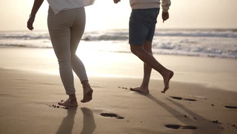 Una-Pareja-Relajada-Caminando-Descalza-Por-La-Playa.-Pasan-Tiempo-Juntos,-Dejando-Huellas-En-La-Arena-Mojada,-Tomados-De-La-Mano.-Suave-Puesta-De-Sol-En-El-Fondo.-Vista-Rara