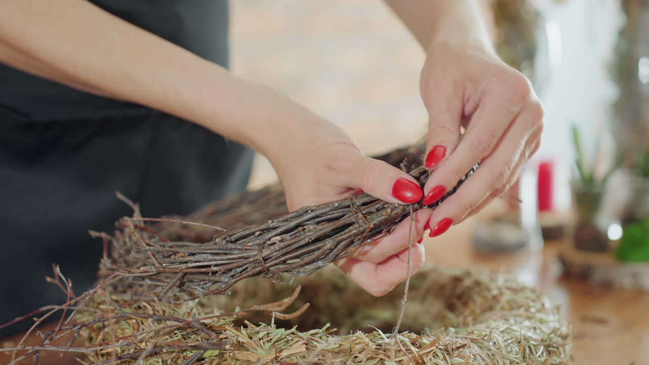 Female hands tying dry twig wreath on straw base using thin wire, focusing on handmade rustic decoration, detailed crafting process, and natural materials for seasonal floral design and home decor