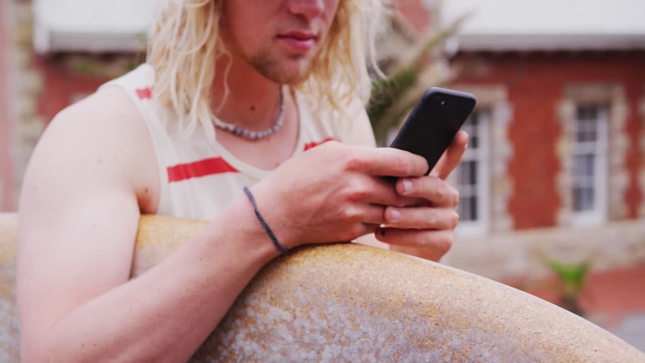 surfista caucásico que sostiene una tabla de surf de madera y usa su teléfono inteligente