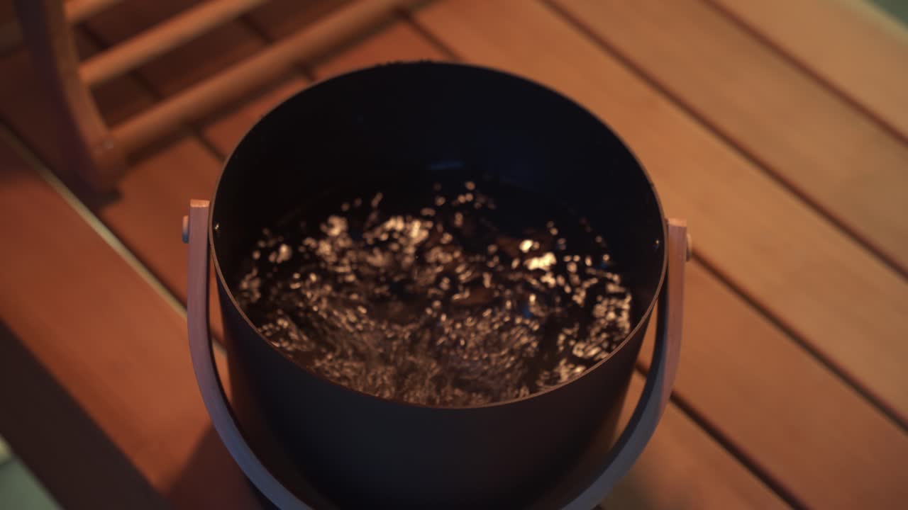 человек, использующий большую металлическую ложку, чтобы собирать воду из роскошного металлического ведра внутри премиальной сауны ночью с оранжевым светом, когда вода брызгает обратно в ведро