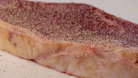 Close-Slow-Motion-Slider-Shot-of-Seasoning-a-Raw-New-York-Strip-Steak-With-Cracked-Black-Pepper