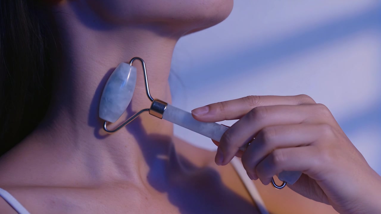 Placing jade roller below jawline, Asian woman massaging neck for skincare in room, thin strap top