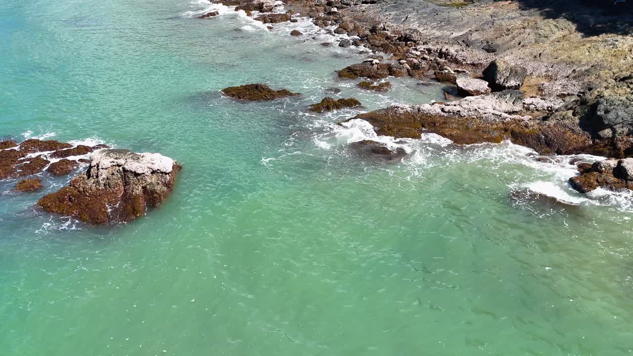 Aerial footage glides above a rocky coastline with turquoise waters and sunlit waves at Port Douglas, Queensland, Australia. Smooth drone movement, bright natural daylight