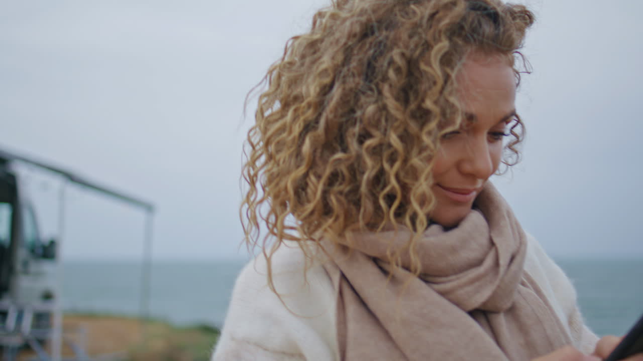una mujer sonriente escribiendo mensajes de texto en el teléfono móvil caminando en un parque de campings en la orilla del mar de cerca.