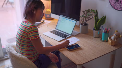 young asian malaysian chinese woman working from home answering a call and taking notes sitting on a desk from the side