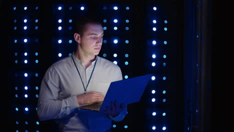 Der-Junge-Mann-Hält-Das-Gerät-In-Der-Hand-Und-Schaut-Auf-Den-Bildschirm,-Inspiziert-Geräte-Oder-Hardware-Rack.-Männlicher-Programmierer,-Der-Mit-Laptop-Arbeitet-Und-Den-Service-Unterstützt,-Während-Er-Im-Rechenzentrum-Spbas-Steht.