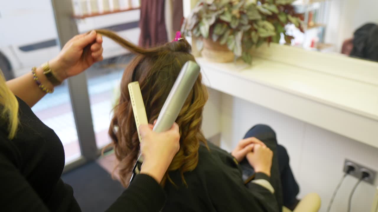 Hairdresser Styling Woman's Long Brown Hair with a Flat Iron in a Salon
