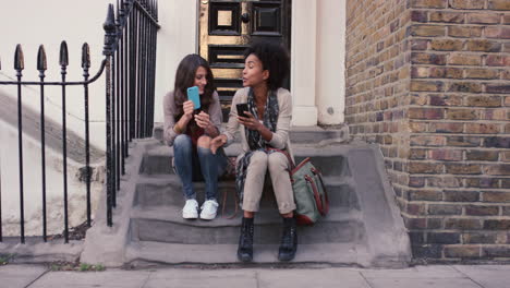 two beautiful woman friends sitting on steps having fun using smart phone