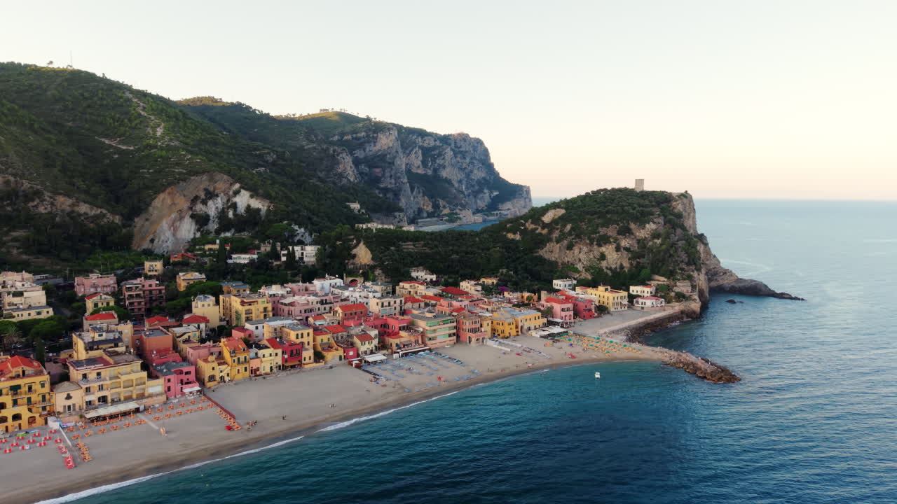 Drone flies forward toward the tip of Varigotti, Liguria. It shows the colorful town, beach, surrounding greenery, and mountains at sunset