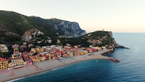 drone flies forward toward the tip of varigotti, liguria. it shows the colorful town, beach, surrounding greenery, and mountains at sunset