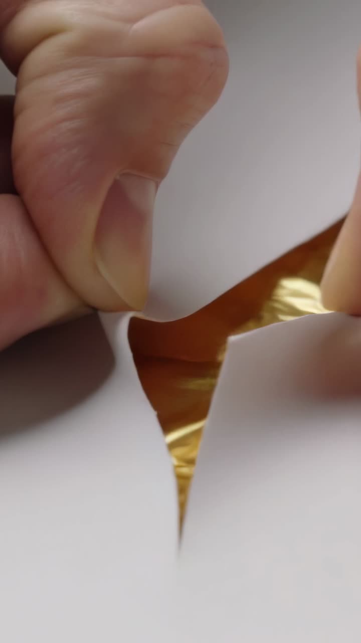 Vertical video: Cutting scalpel piercing paper on table, hands peeling gold foil revealing layer