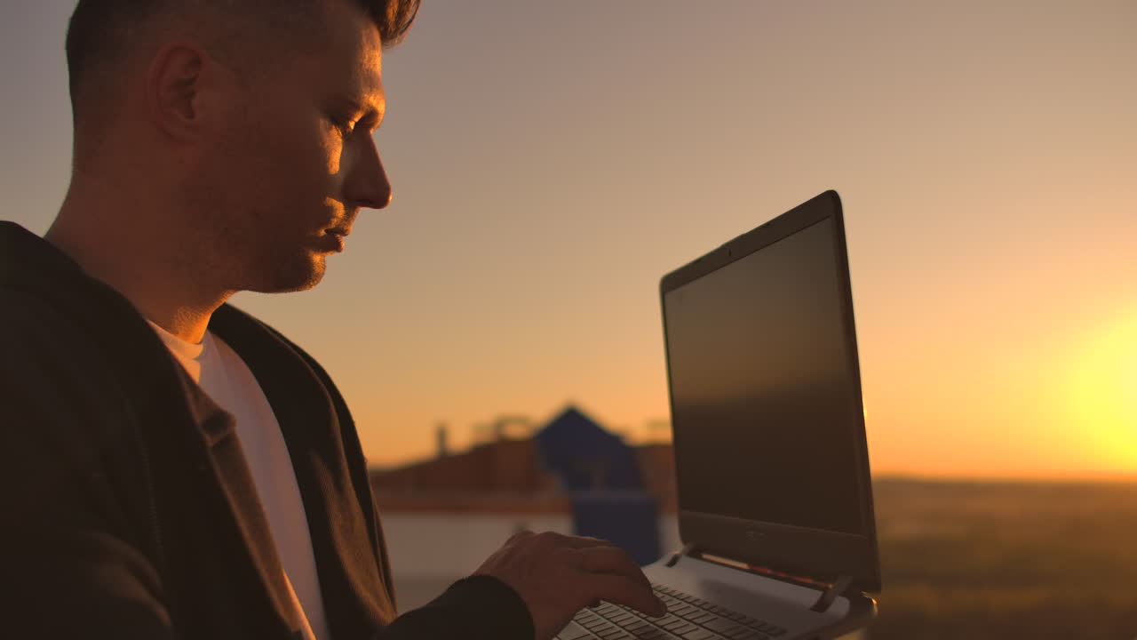 Hacker using laptop on rooftop with city view and forex chart. Hacking and stats concept. A man at sunset in slow motion writing software code on a laptop
