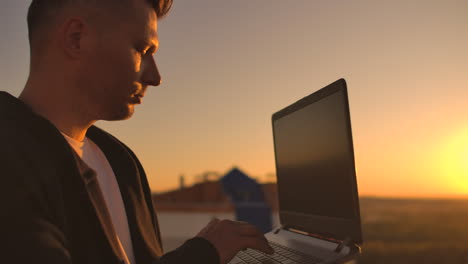 hacker using laptop on rooftop with city view and forex chart. hacking and stats concept. a man at sunset in slow motion writing software code on a laptop