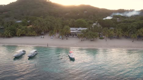 cinematic aerial view of caribbean coast with boats, turquoise water, white sand beach, resorts at sunrise with island background, roatan island,west end, honduras