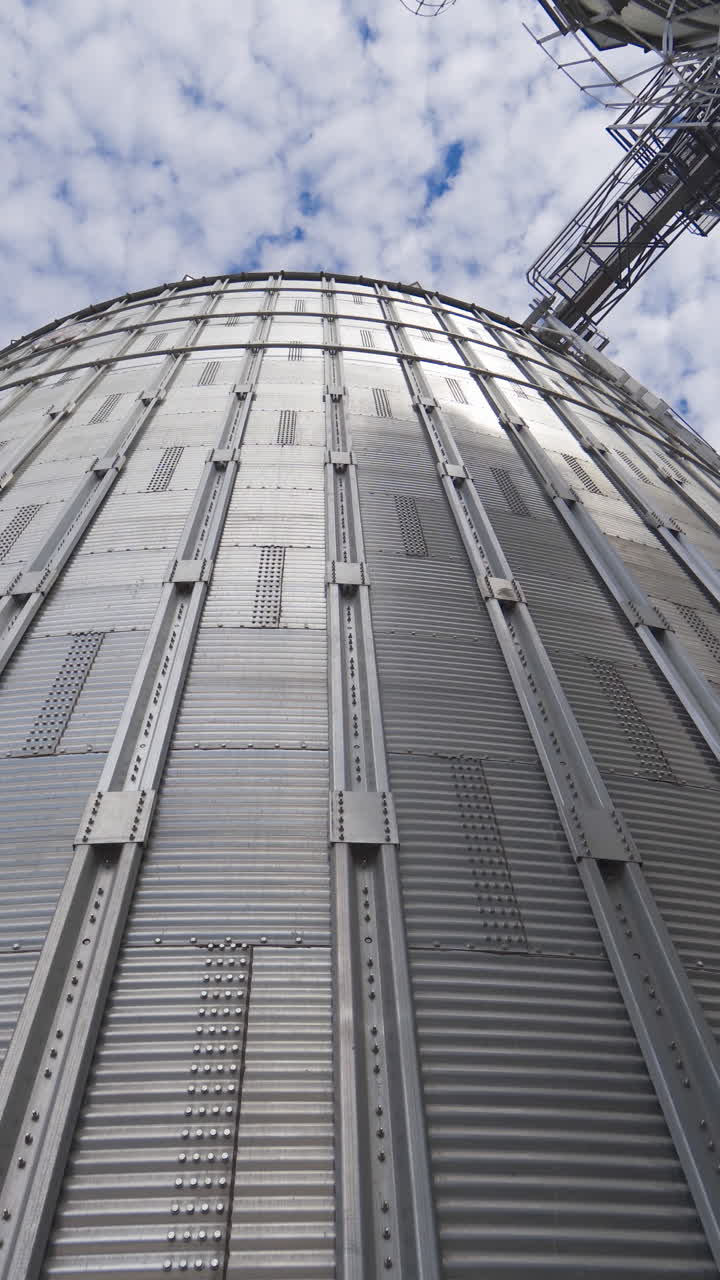 Large metal grain elevator outdoors. Modern silver granary on sky background. Huge aluminum container for storing crop. View from below. Vertical video