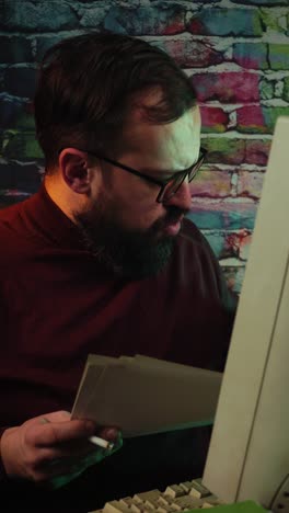 man from 1980's in front of desktop computer, smoking cigarette and looking at paper documents