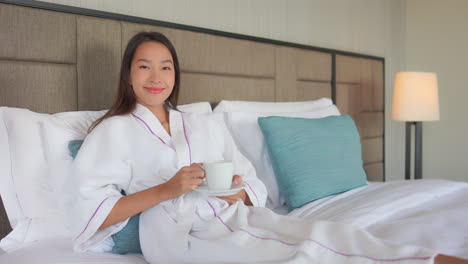 Young-asian-woman-lying-on-a-hotel-bed,-wearing-white-dressing-gown-and-holding-a-cup-of-coffee-or-tea,-looking-relaxed-and-smiling