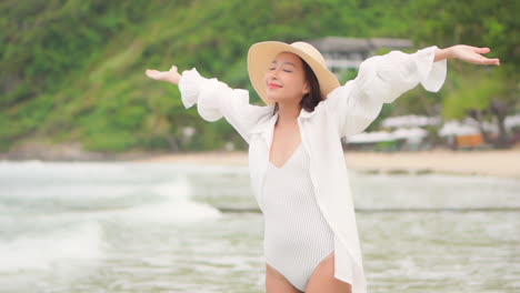 Bella-Dama-Asiática-Con-Traje-De-Baño-Blanco-Y-Blusa-Extendiéndose-Y-Levantando-Los-Brazos-Sonriendo-Durante-Un-Paseo-Matutino-En-La-Playa-De-La-Isla,-Mareas-Marinas-Y-Montañas-Verdes-En-El-Fondo,-A-Cámara-Lenta