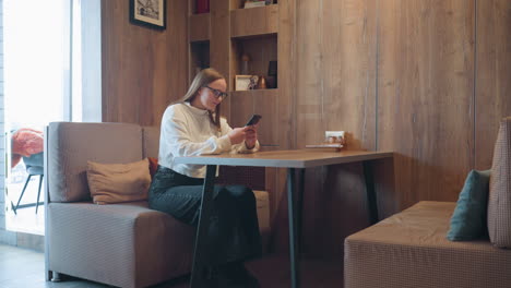 woman in white sweater seated on cushioned bench at wooden table chatting over phone in cozy cafe setting with warm lighting, books on shelf, and notepad on table