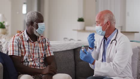 Senior-caucasian-male-doctor-preparing-vaccination-for-senior-man-at-home-both-wear-face-masks