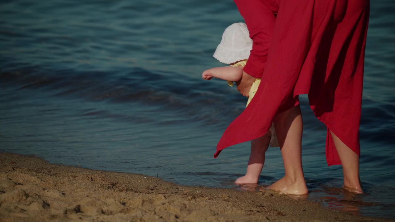 Madre che tiene in braccio il bambino, aiutando i piedini a toccare l'acqua del mare e la sabbia per una prima esperienza in riva all'oceano. Momento di vacanza estiva in famiglia