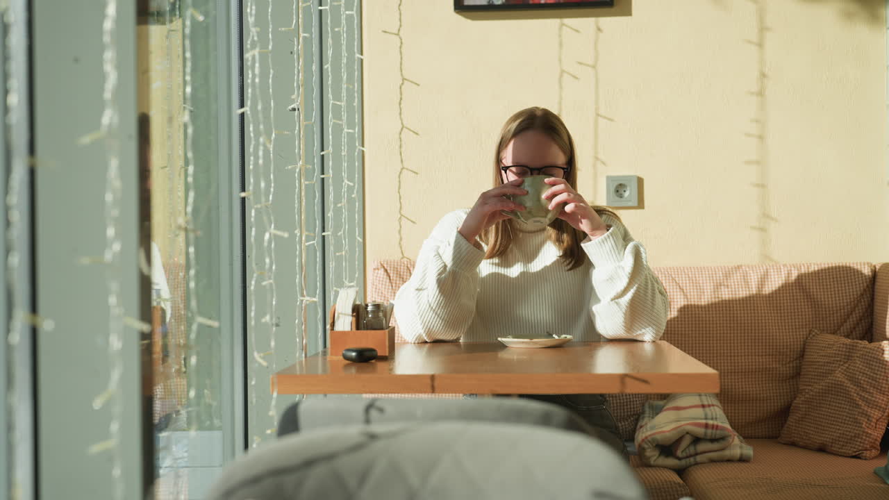 Lady in white sweater enjoys tea while seated on cushioned bench in cozy cafe, decorative string light reflections cast soft patterns on warm colored wall