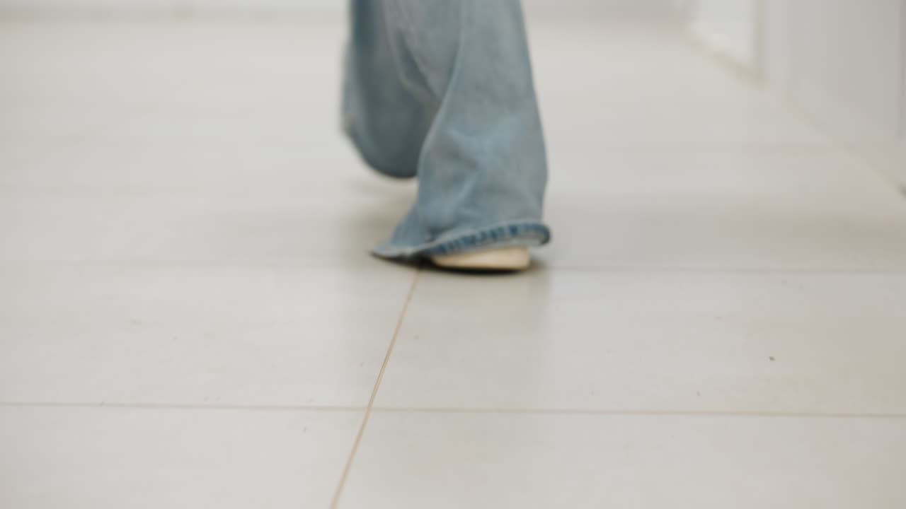 Back leg view of woman in jeans and white sneakers walking along bright corridor, low angle on tiled floor, soft blur, business owner showing purposeful stride in indoor laundromat, calm ambiance