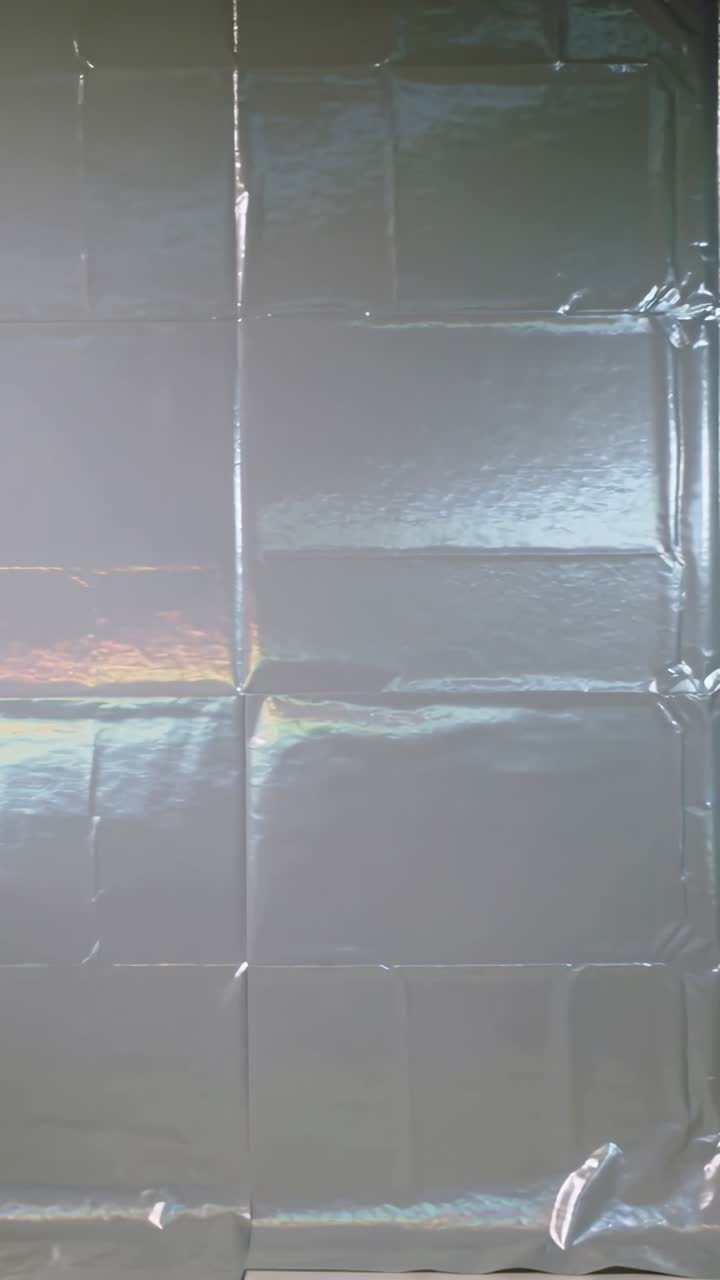 Video verticale: Luce che si sposta su una pellicola iridescente in primo piano, rivelando riflessi e pieghe.