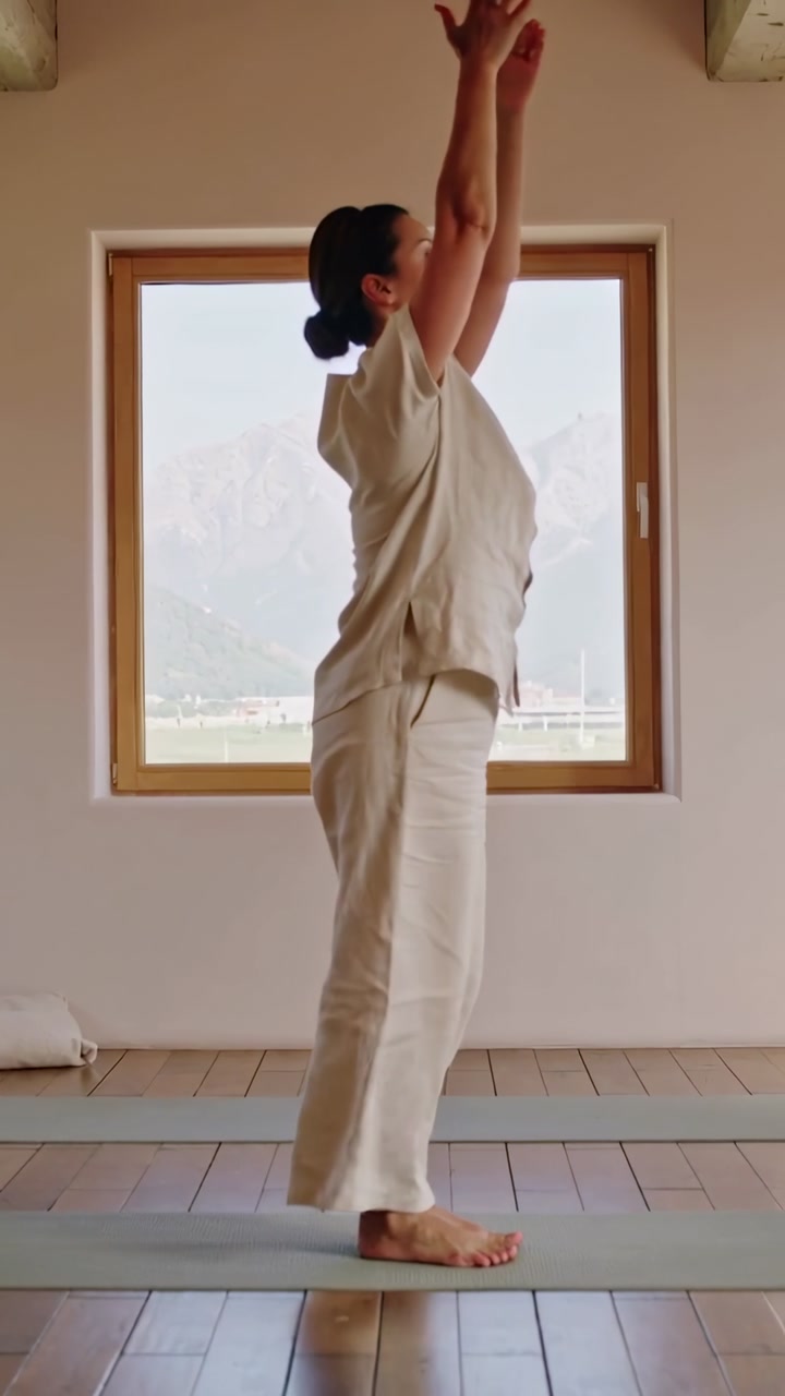 Vertical video: Breath cueing woman rising, folding forward on mat in room warming in beige clothes