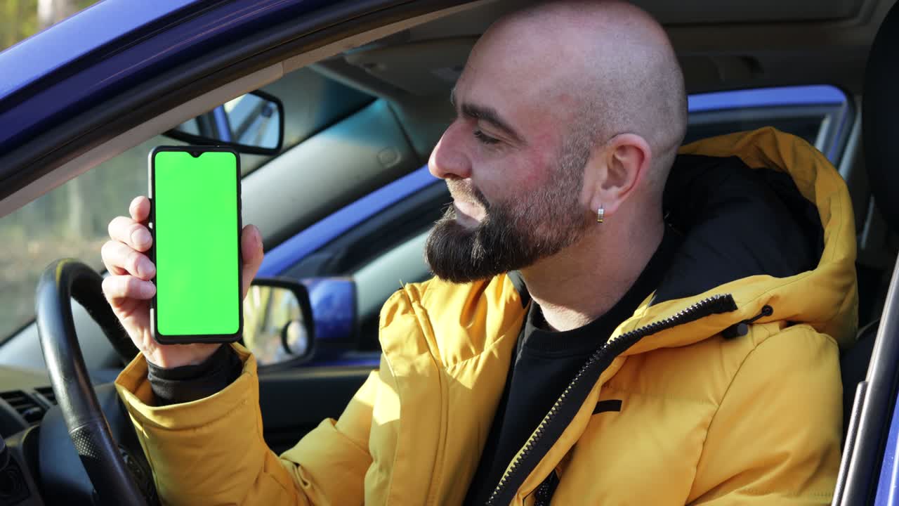 retrato de un hombre barbudo sentado en el coche azul, sonriendo y mostrando un teléfono móvil con pantalla verde. llave de croma