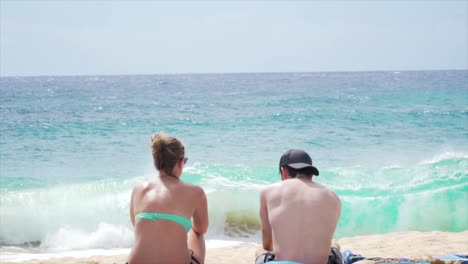 couple sitting in front of ocean talking, as waves crash in front of them in slow motion