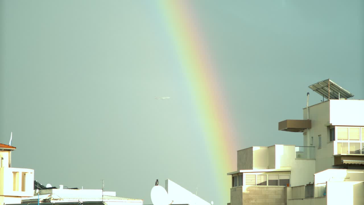 una toma panorámica de un avión volando, luces de arco iris sobre los edificios de la ciudad de israel, nubes deslizantes, cielo azul oscuro, luz del sol en los techos, clima lluvioso, oriente medio tel aviv, sony 4k video