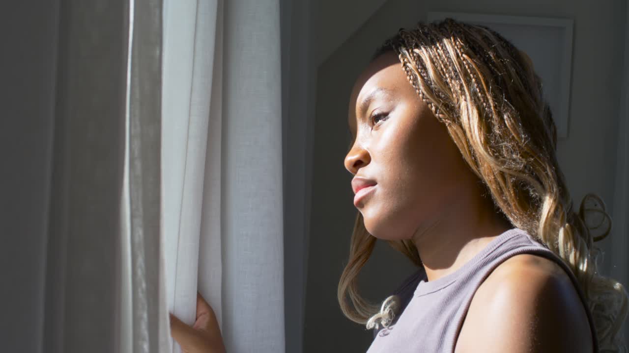 Notando il sole, una donna afroamericana con le trecce tiene la tenda, inclinando la testa e guardando fuori dalla finestra.