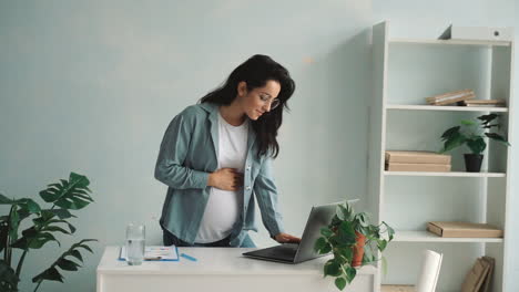 young pregnant business woman works using a laptop massaging and stroking her belly at home