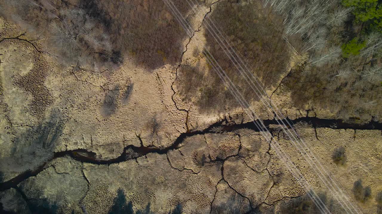 Top down aerial view slowly moving above dark colored narrow Pääsküla river during a spring or autumn sunny day, overgrown brown grass and moss around. Electrical lines are visible on top of nature.