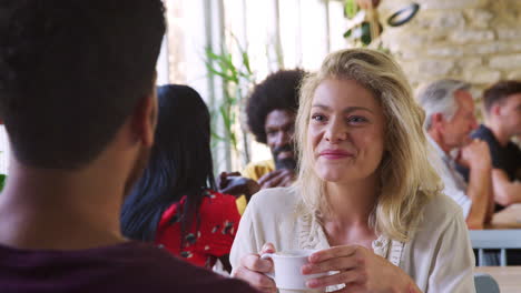 happy young adult woman drinking coffee and talking to her male friend at a table in restaurant, daytime, over shoulder view