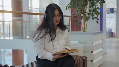 focused young woman wearing headphones and glasses sits on wooden bench in modern mall reading vintage book thoughtfully with blurred background