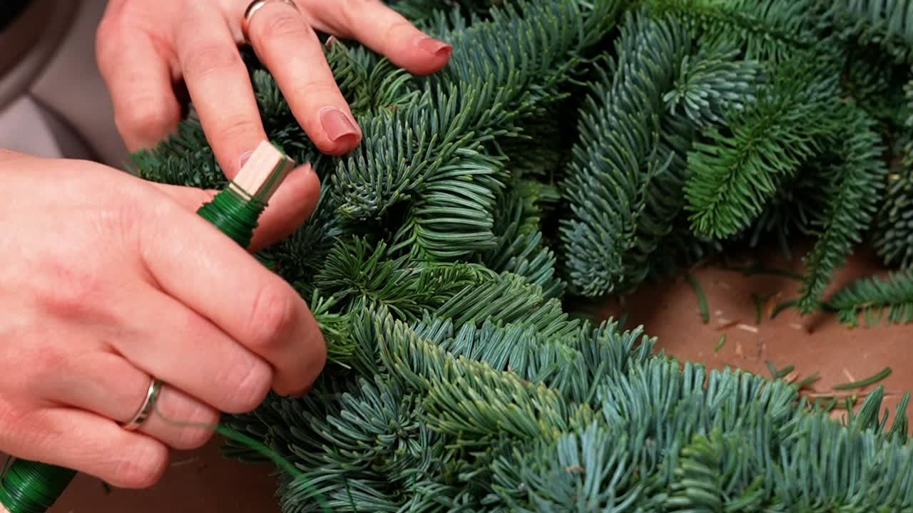 Dettagliata inquadratura ravvicinata di mani che realizzano una bellissima ghirlanda natalizia utilizzando rigogliosi rami sempreverdi. La scena cattura il lavoro intricato, le vivaci tonalità di verde e l'atmosfera festosa.