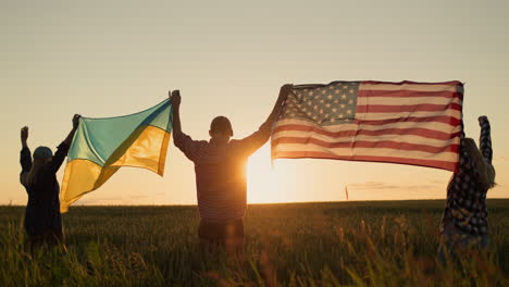 Junge-Menschen-Hissen-Die-Amerikanische-Und-Die-Ukrainische-Flagge-Vor-Dem-Hintergrund-Eines-Weizenfeldes,-Auf-Dem-Die-Sonne-Aufgeht